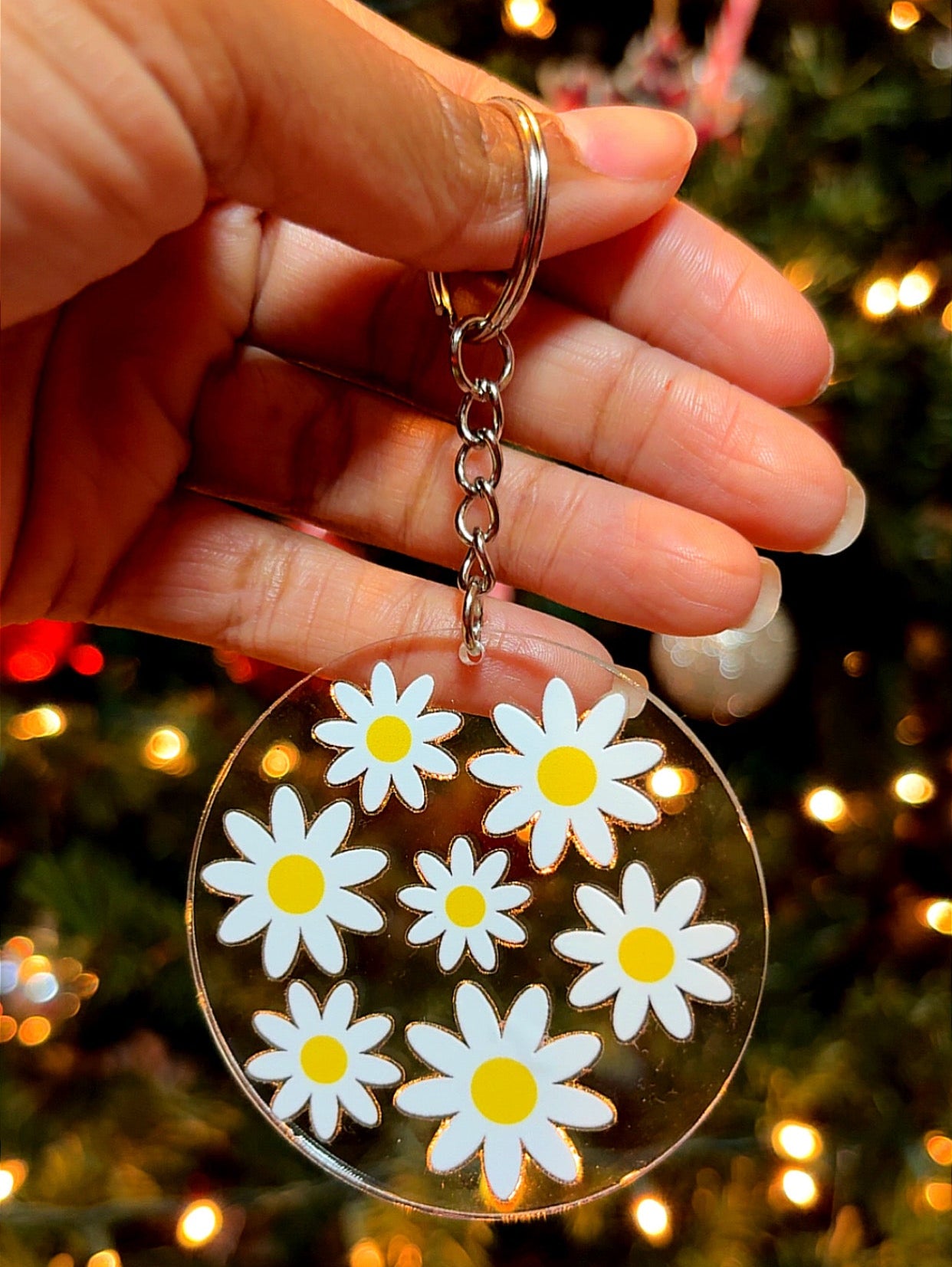 Hand holding a keychain with daisy flower design against a Christmas tree background